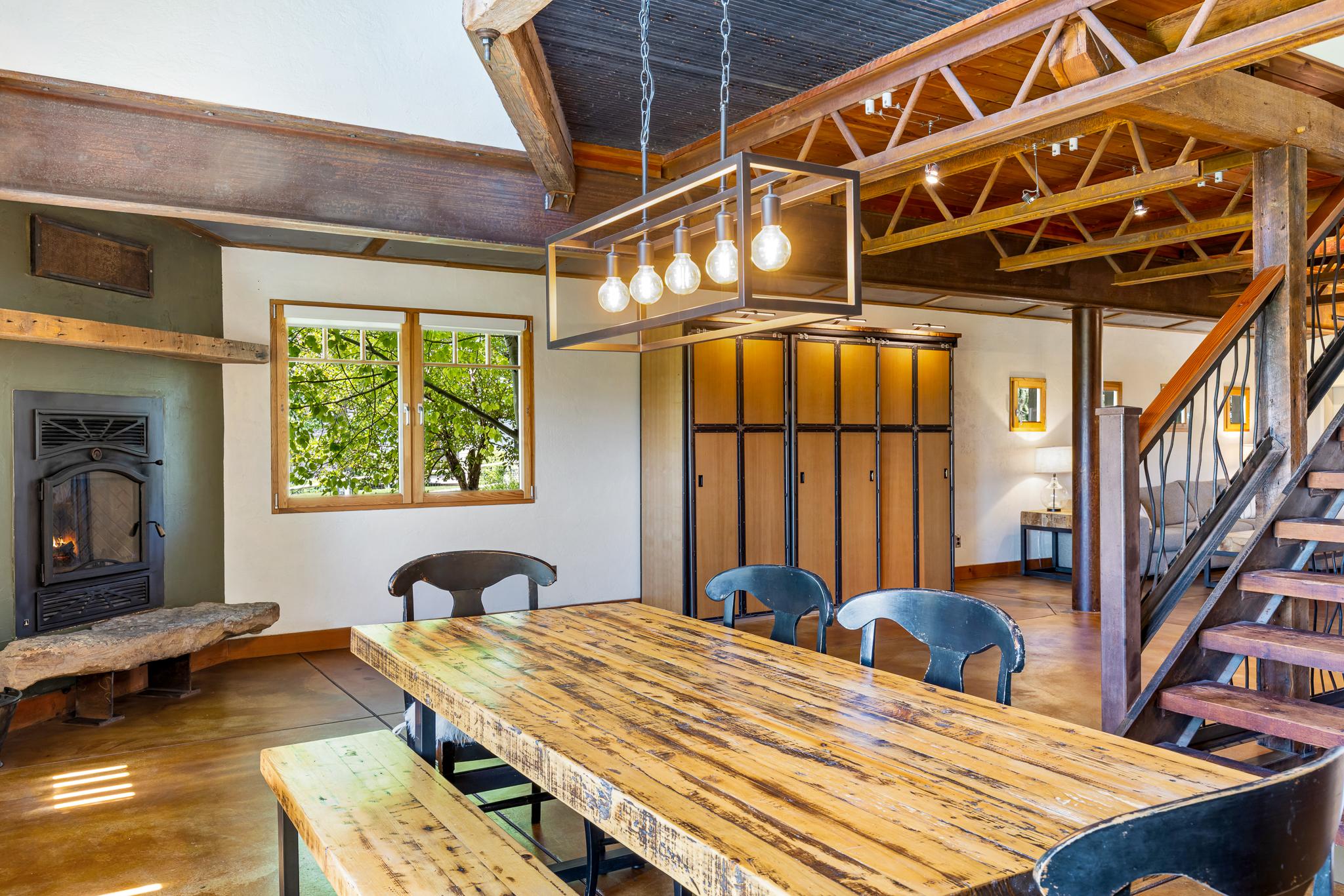 Dining area with reclaimed wood table, Norwegian dual-opening windows, exposed iron trestle beams, and ski lift cable railings in custom timber frame home at 2449 Turkey Red Lane, Bozeman MT.